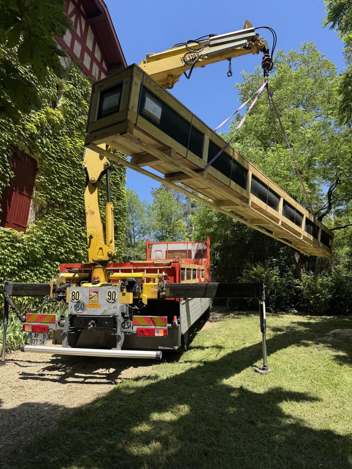 Camion-grue soulevant un long élément conditionné au bord d’un jardin.