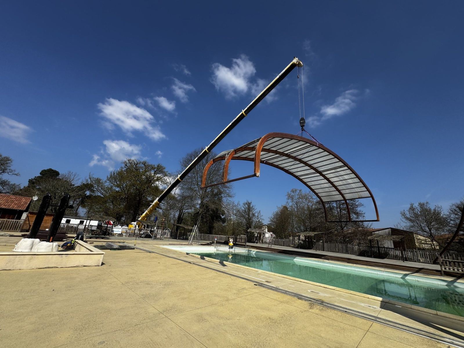 Une grue soulève une travée de l’abri cintré au-dessus de la piscine.